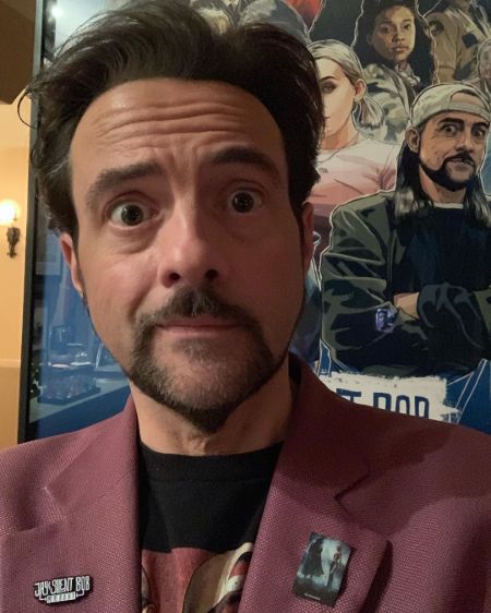 Actor Kevin Smith in a red coat poses for a picture in front of his poster.